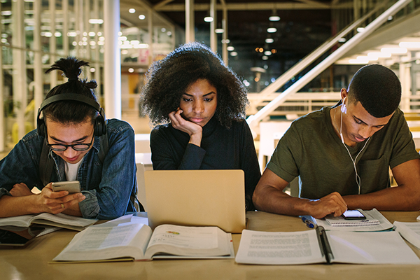 Three graduate students doing research with personal devices and books. 