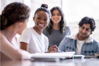 A group of multicultural students sit around a table with open textbooks. An African-American student is the only person in focus. She is smiling at another student in the group.